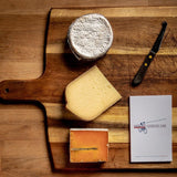 saxelby cheesemongers cheese of the month club - a selection of three cheeses on a cheese board with a cheese knife and cheese journal